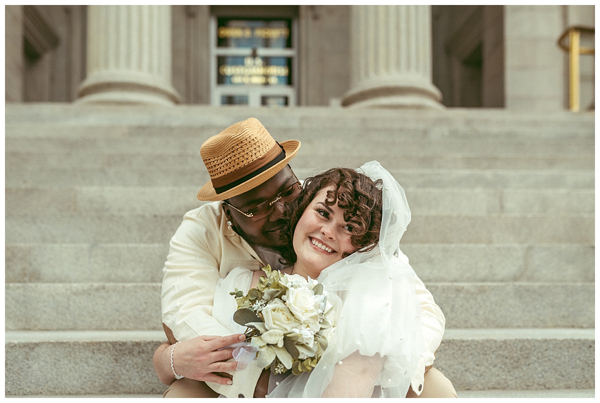 man hugs woman while sitting on steps at wedding portrait session