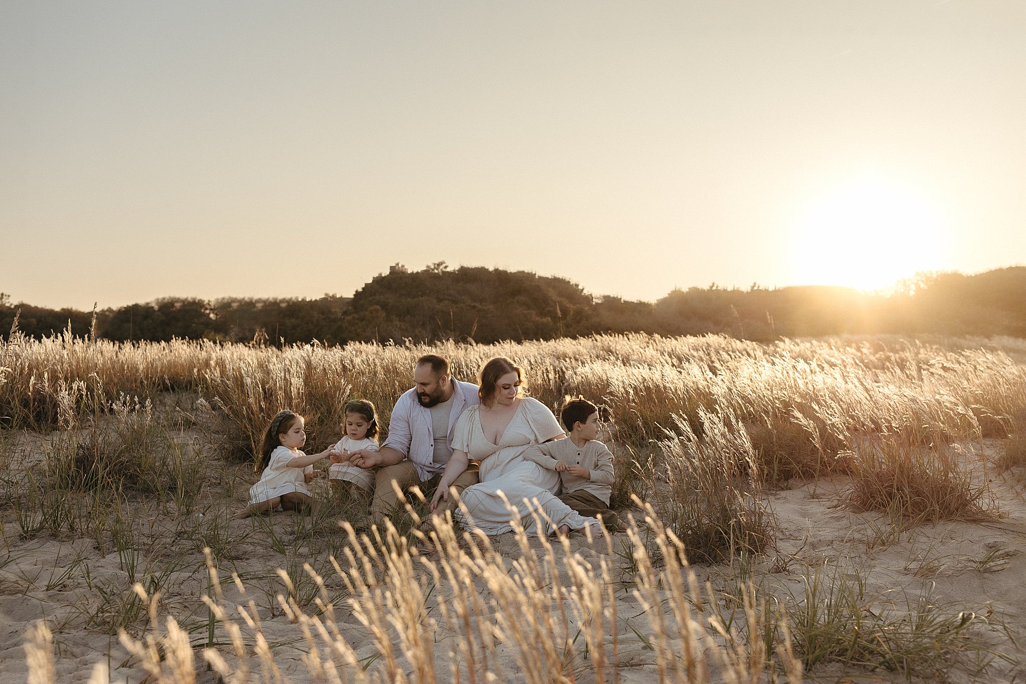 parents sit with kids on sand as sun sets behind them at perfect family session