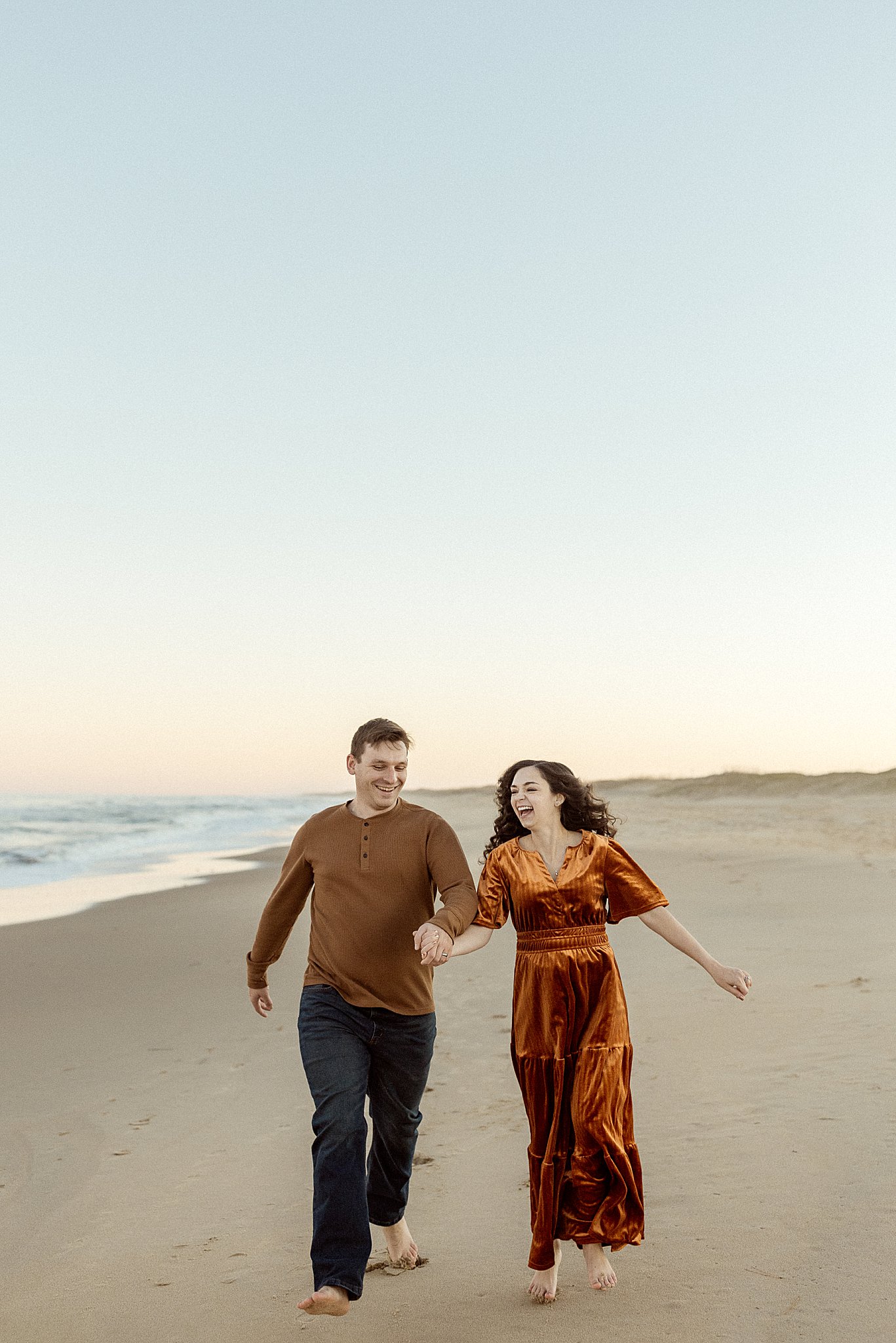 couple laughs as they walk along sand holding hands by Virginia Beach Photographer 