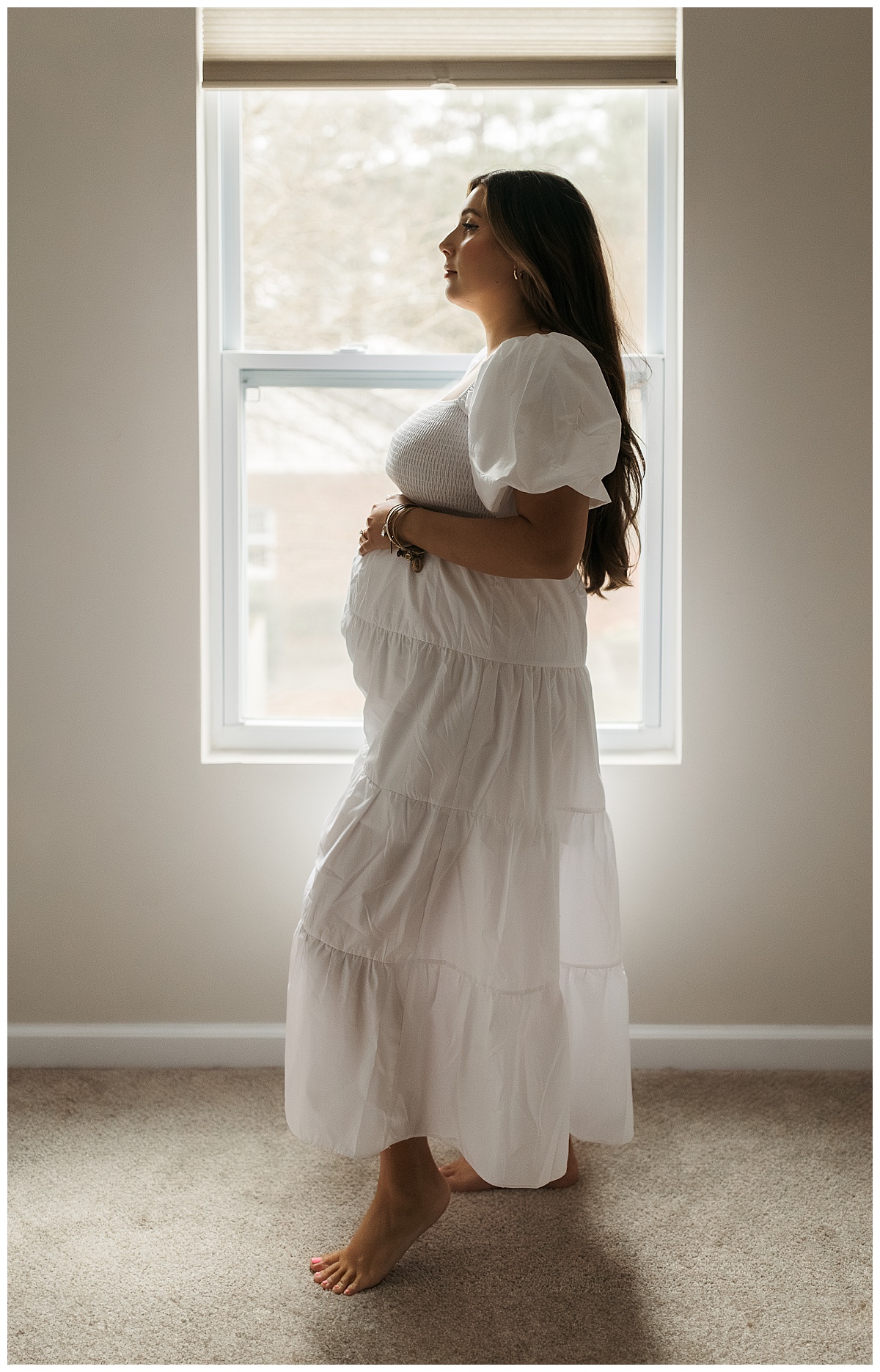 mom-to-be holds her pregnant belly as she stands in front of a window by Nikki Meer Photography