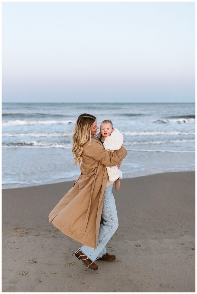 mom walks along sand holding toddler wearing a long coat during beach family session